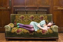 A woman wearing purple tights and a blue dress naps on a green velvet sofa.