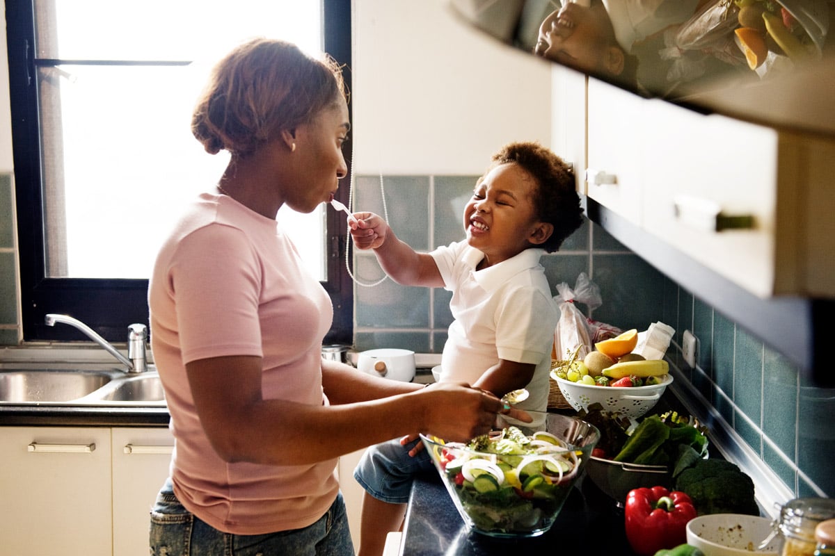 A woman makes food in her kitchen as her toddler feeds her. 