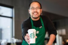 A barista smiles as they present a pumpkin spice latte in a to go cup at Starbucks.
