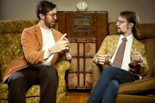 Two men argue while holding an adult beverage in yellow chairs.