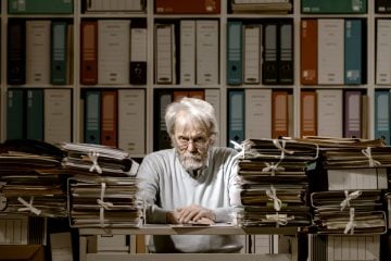 A man sits at a desk with paperwork surrounding him meant to represent his debt.