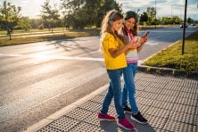 Two girls look at their phones while wearing headphones and walking outside.