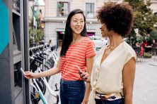 Two women use an ATM outdoors. One is holding her debit card while the other is holding her cell phone. They are conversing with one another.