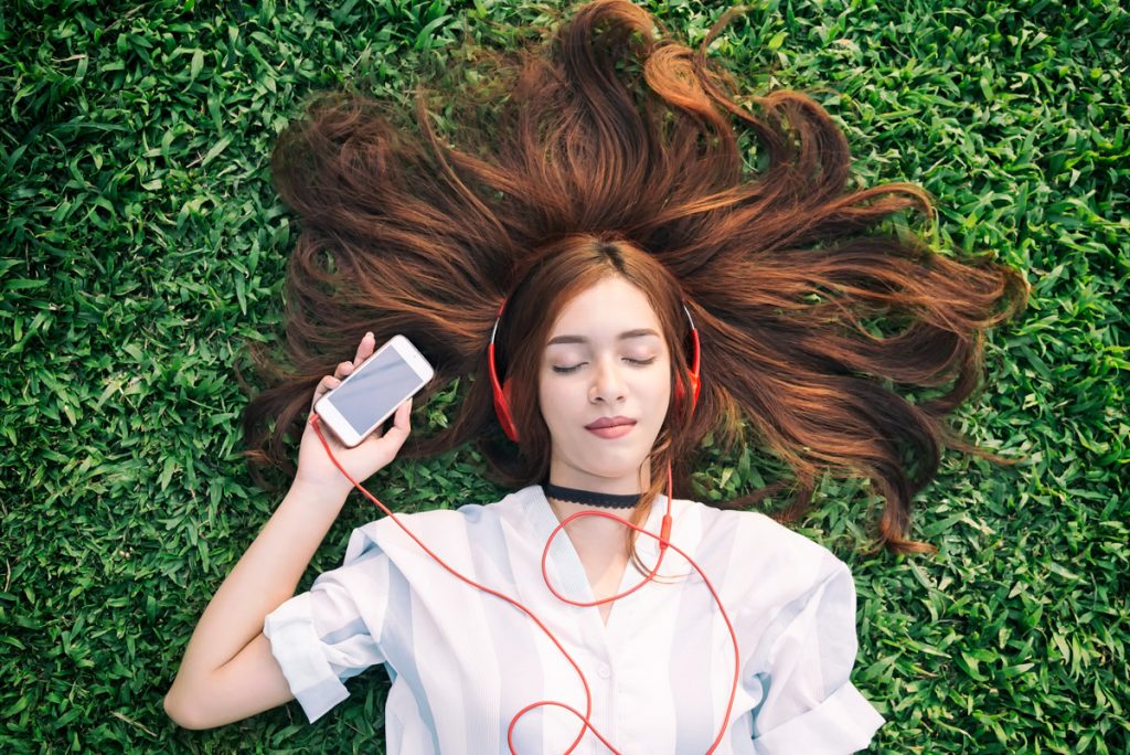 A woman lays in grass with a peaceful expression on her face while listening to an audio book.