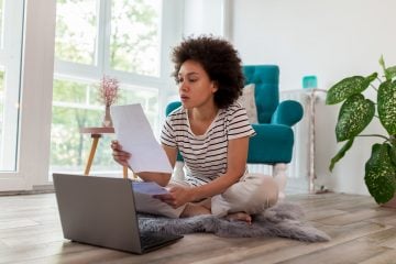A woman looks over her bills while sitting on the floor with her laptop.
