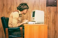 A man types typically on an old school computer. The man looks like he&rsquo;s from the 1970s or 1980s.