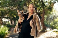 A woman wears a black dress, pears and a fur coat. She&rsquo;s also holding a small dog.