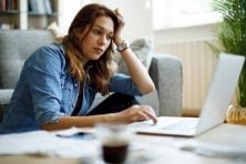 A woman looks stressed as she looks at her computer while sitting on the floor in her living room.