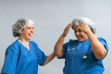 Two nurses wear hair nets and scrubs while laughing.