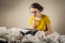 A woman ponders what to write about on her type writer with crumbled up pieces of paper all around her.