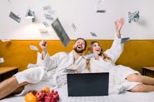 A couple throw money in the air while in their bed with robes on.