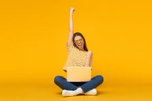 A young woman celebrates. She is sitting against a yellow background.