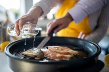 A couple pour a glass of wine into the pan they are cooking meat and fish with.