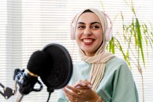 A woman wearing a hijab smiles as she records something into a microphone at her home office.