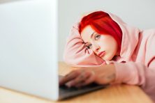 A woman waits for her internet to load. She looks bored. She has red hair and is wearing a pink hoodie.
