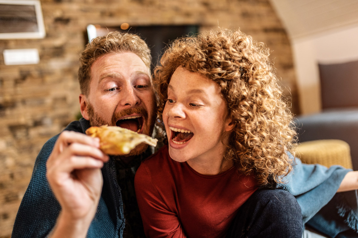 Two people with red hair both go to take a bite of the same slice of pizza.