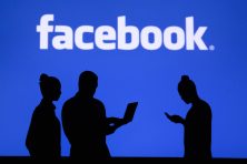 People look at their laptop and phone with the Facebook logo behind them. The people are silhouetted.