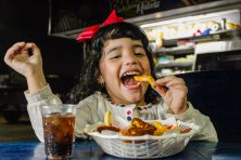 A little girl joyously bites into a French fry with cheese on it in a diner.