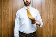 A man holds a cup of coffee with his tie dipped into the coffee.