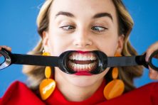 A woman uses salad tongues to open up her mouth to show her teeth.