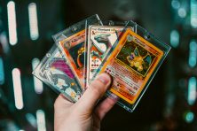 A hand holds four Pokemon cards in protective plastic sleeves.