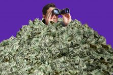 A man peeps out of a tall stack of money with binoculars.
