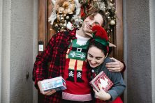 A man and woman hug outside of a home while wearing Christmas themed outfits.