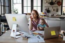 A woman looks stressed while looking at finances on her home computer with her baby in her lap.