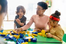 A daycare provider helps two kids build a structure with blocks.