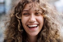 A woman with curly hair laughs while looking into the camera.
