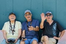 Three retired men laugh as they hold remote controllers outside on a bench.
