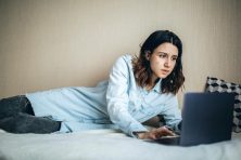 A woman lays down on her bed while doing paperwork on her computer.