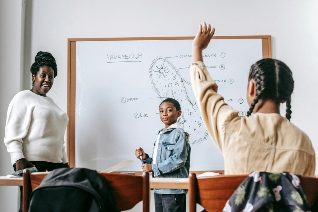 A student raises her hand in class with her teacher looking at her and another student at the white board. 