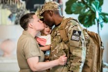 A mother holds her toddler as she kisses her husband in a military uniform inside their home.