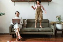 A woman works from home with her daughter jumping on the couch.