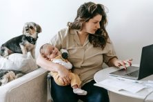 A woman works from home with her newborn in her arms and her two dogs behind her on the couch.