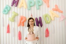 A lady wearing a birthday hat with a happy birthday banner in the background, holds a pink birthday cake