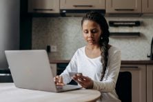 A woman looks at the back of her credit card while she types on her laptop.