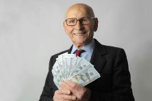An older gentleman in a suit and tie holds a fan of $100 bills while smiling.