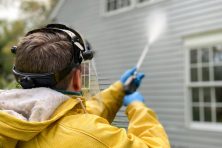 A man dressed in a raincoat pressure washes a house.