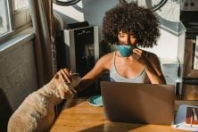 A woman sits at her kitchen table sipping coffee, checks her computer, and pets her dog while sunlight beams through the window.