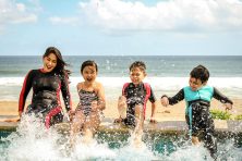 A family splashes in a pool with the ocean behind them while on summer vacation.