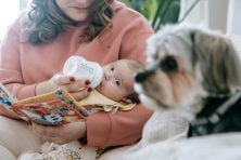 A mother feeds her baby a bottle while the baby is looking at their dog.