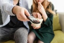 One person holds a TV remote while another eats a snack.