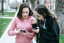 Two young girls look at their cellphones together.