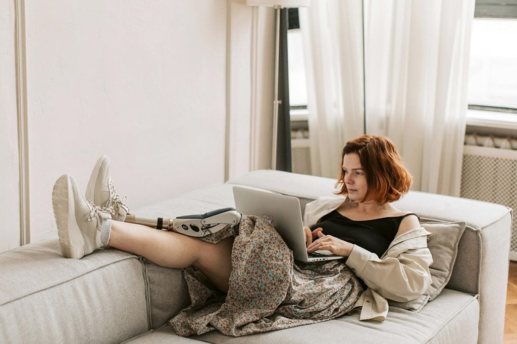 A woman works from her laptop while lounging on the couch. He has a prosthetic leg.