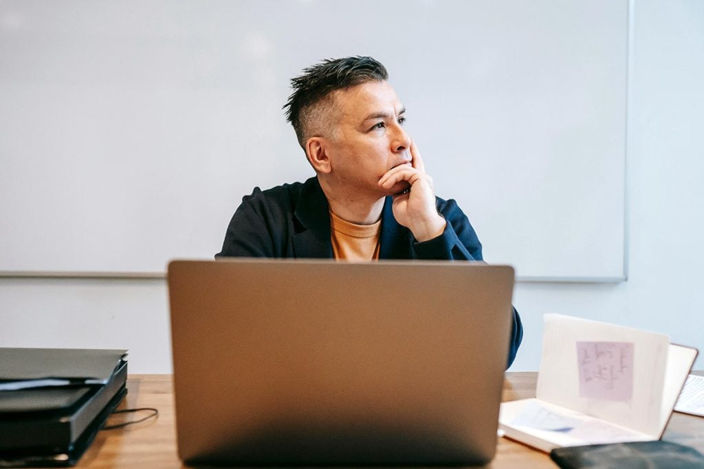 A man looks out a window while sitting near a laptop pondering a question.
